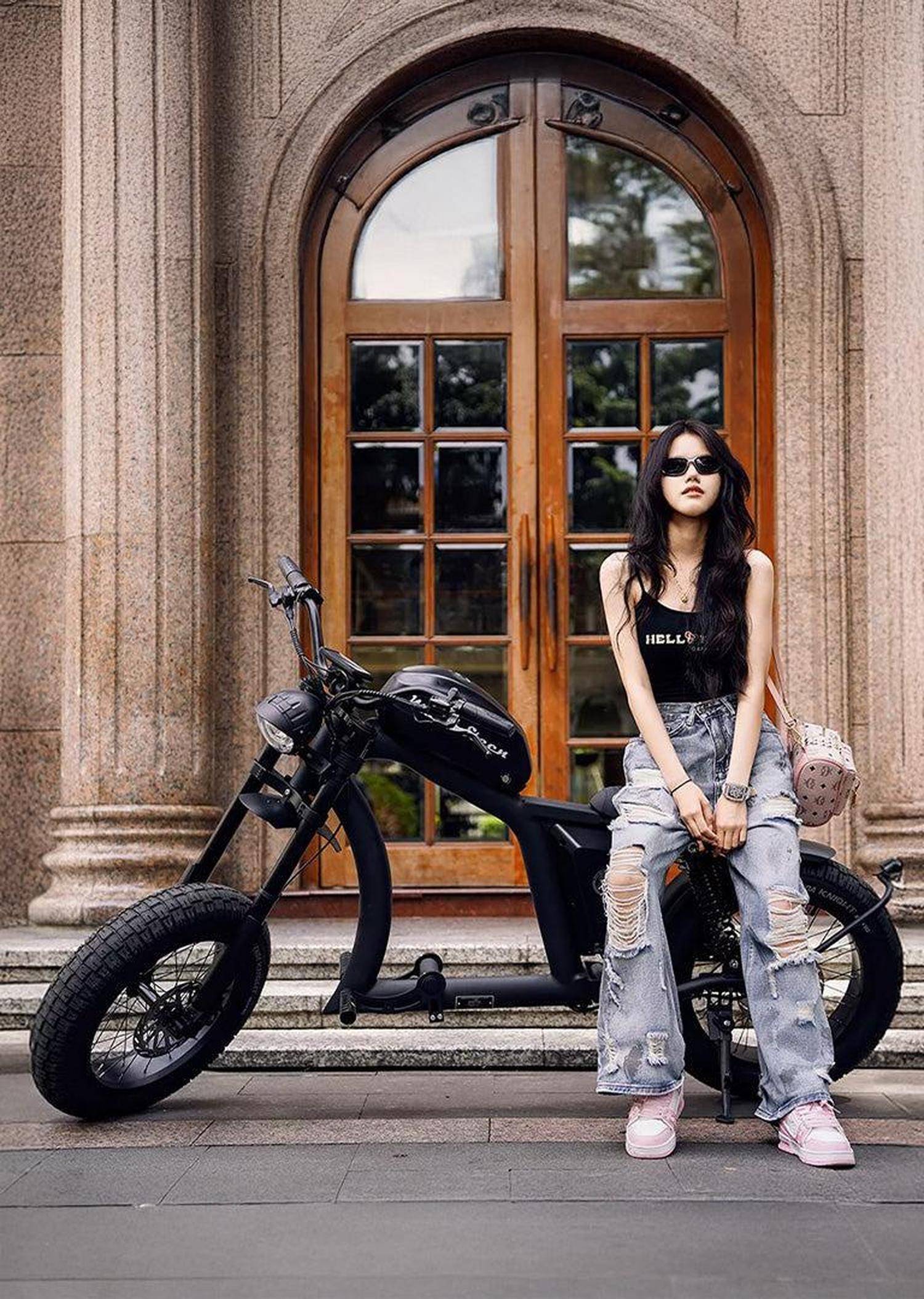 Woman posing with a black scooter in front of a large wooden door.