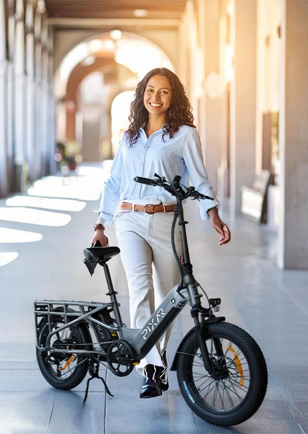 Woman holding a black electric bike in an outdoor setting with sunlight filtering through arches.