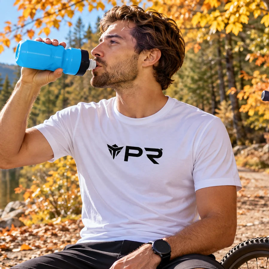 Rider in a white Punk Ride T-shirt drinking from a blue cycling bottle beside an autumn lake.