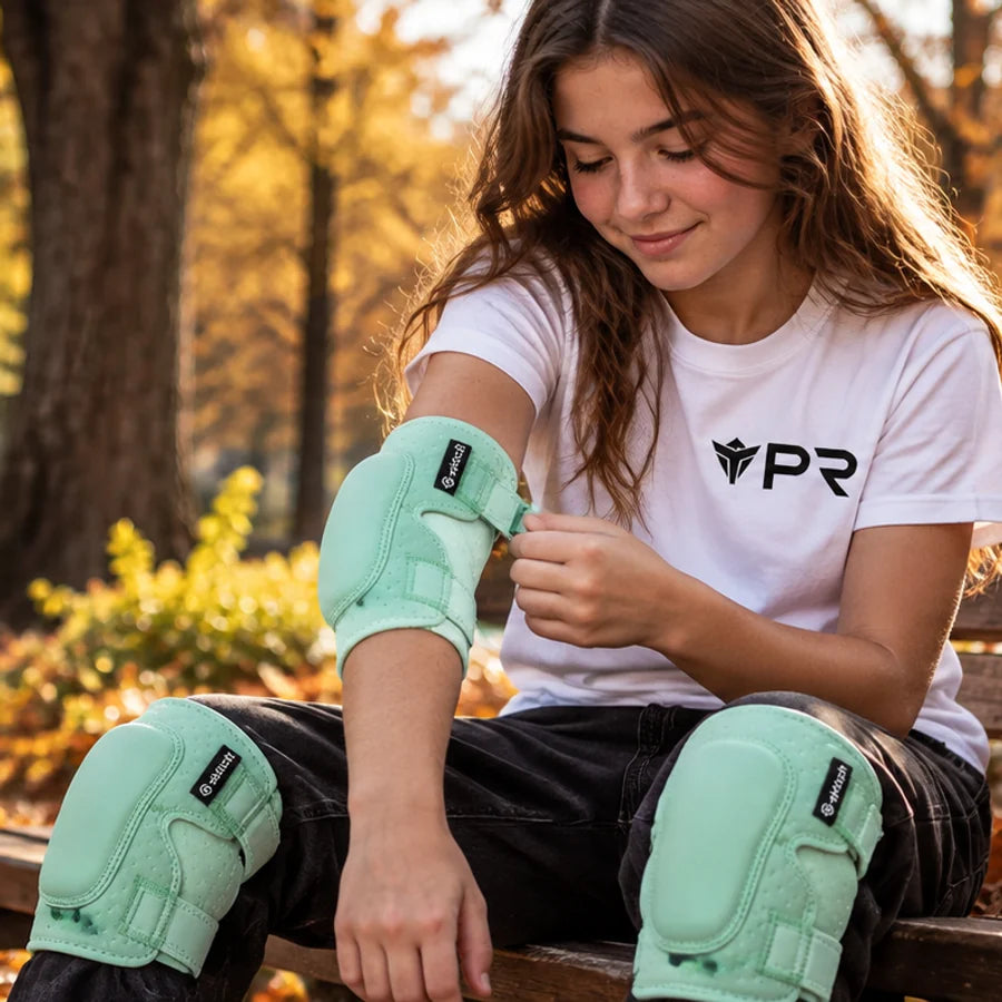 Young rider in a white Punk Ride T-shirt adjusting mint-green protective pads on an autumn park bench.