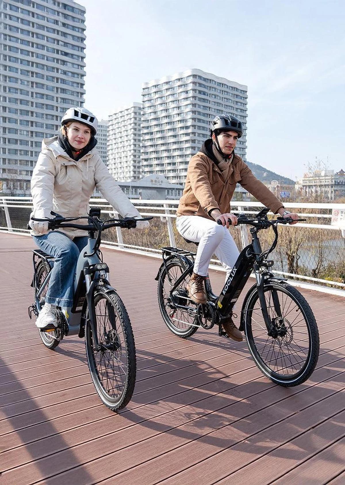 Two people riding electric bikes on a bridge with buildings in the background