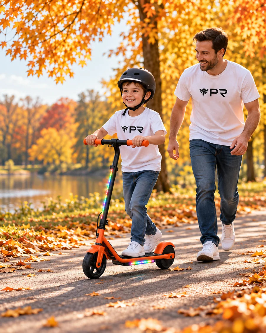 A helmeted child rides an orange iScooter K2 Pro beside an adult in white Punk Ride logo T-shirts on an autumn lakeside path.